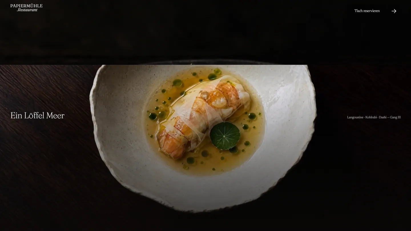 Gang „Ein Löffel Meer“ auf papiermühle-restaurant.de: eine Langoustine in hellem Dashi mit grünen Kräuterölperlen auf einem weißen, unregelmäßig geformten Teller vor schwarzem Grund, links der Name des Gangs, rechts die Zutaten